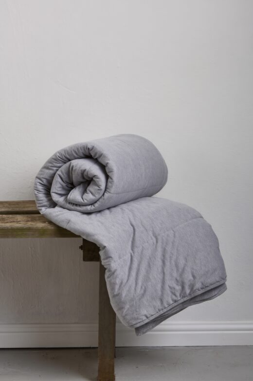 A rolled grey melange quilt resting on a rustic wooden bench against a plain white wall.